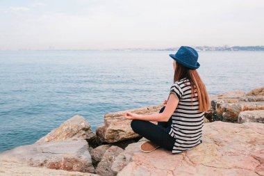 Şapkalı yoga uygulamak ve denizin yanında taşlar üzerinde meditasyon genç güzel kız. Rekreasyon. Tatil. Gevşeme.