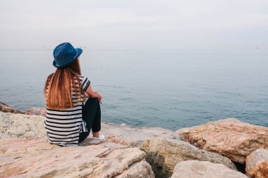 Şapkalı genç güzel kız kayalar sahil üzerinde oturur ve mesafe içine görünüyor. Dinlenme, tatil, dinlenme.