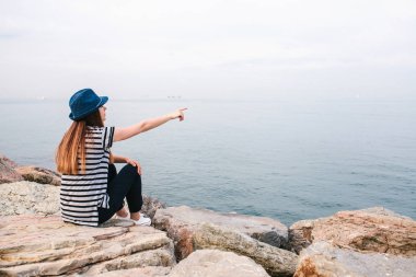 Şapkalı genç güzel kız kayalar deniz kenarında oturur ve elini mesafe gösterir. Dinlenme, tatil, dinlenme.