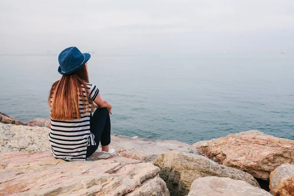 Junge schöne Mädchen mit Hut sitzt auf den Felsen am Ufer des Meeres und blickt in die Ferne. Ruhe, Urlaub, Entspannung. — Stockfoto