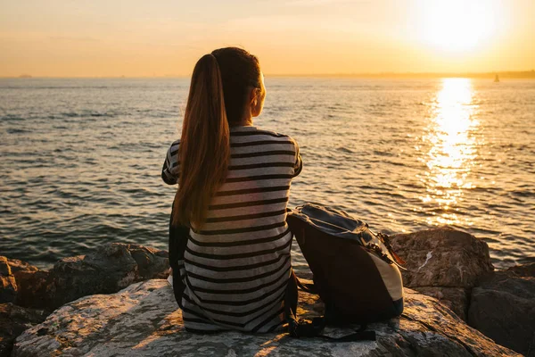 Ein junges Touristenmädchen mit Rucksack sitzt bei Sonnenuntergang auf den Felsen am Meer und blickt in die Ferne. Erholung, Entspannung, Reisen, Urlaub. — Stockfoto