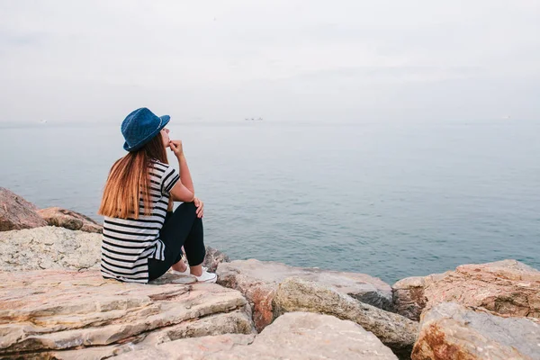 Junge schöne Mädchen mit Hut sitzt auf den Felsen am Ufer des Meeres und blickt in die Ferne. Ruhe, Urlaub, Entspannung. — Stockfoto