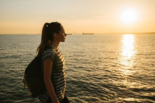 Una joven turista con una mochila se para junto al mar al atardecer y mira a la distancia. Descanso, relajación, viajes, vacaciones . — Foto de Stock