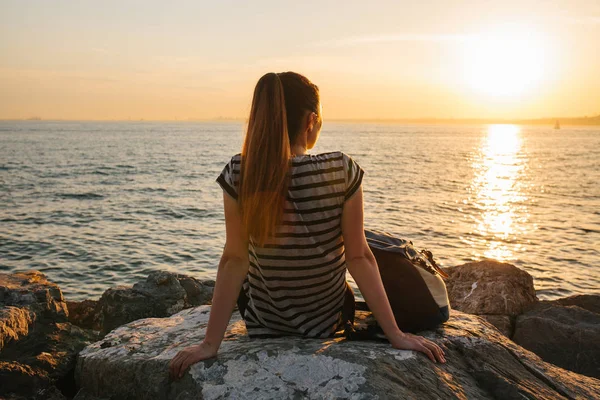 Ein junges Touristenmädchen mit Rucksack sitzt bei Sonnenuntergang auf den Felsen am Meer und blickt in die Ferne. Erholung, Entspannung, Reisen, Urlaub. — Stockfoto