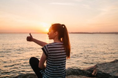 Genç bir kız günbatımında kayalar denizin yanında oturur ve serin anlamına gelir bir el işareti gösterir. Dinlenme, tatil, dinlenme.