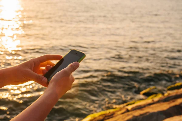 Las manos femeninas sostienen el teléfono. Utilice el teléfono para conectarse en el viaje. Concepto de viaje. Vista del mar y puesta de sol en el fondo . — Foto de Stock