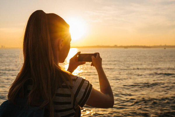 A young girl with a backpack pictures on the phone a beautiful view of the sea and the sunset. Picture for memory. Travel, vacation.
