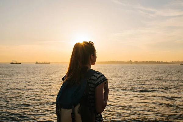 Una joven turista con una mochila se para junto al mar al atardecer y mira a la distancia. Descanso, relajación, viajes, vacaciones . — Foto de Stock