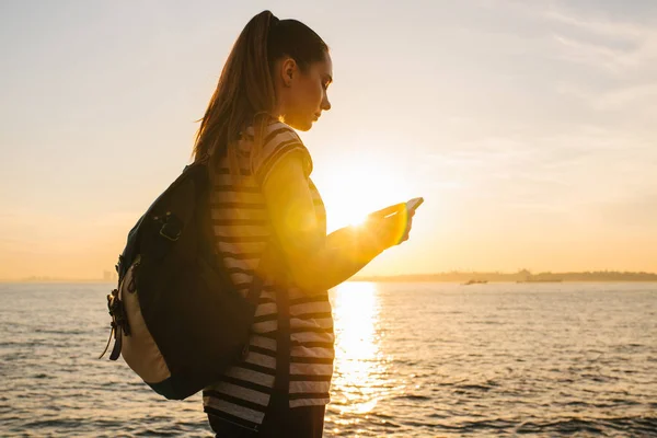 Joven hermosa chica con una mochila utiliza un teléfono para conectar. Hermosa vista del mar y puesta de sol en el fondo . — Foto de Stock