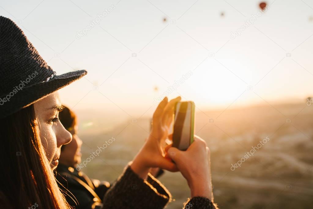 Side view of charming brunette girl in hat takes picture on smartphone during flights of lots of hot air balloons on warm sunny day.