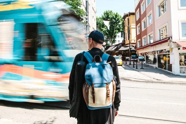 Muž stojící u silnice s tramvají. Mužské turista v neformálním oblečení s batohem stojí před tramvají silnice na slunečný letní den. Rozmazání pohybu. — Stock fotografie