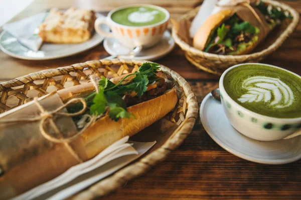 Здоровый завтрак в ресторане: чашка зеленого чая с молоком, размытый десерт и два сэндвича с овощами и травами на деревянном столе . — стоковое фото