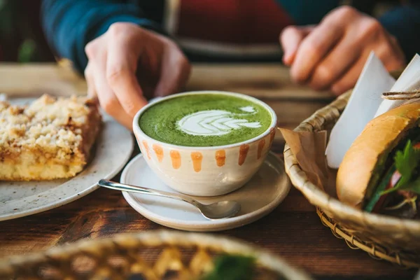 Крупным планом человек руки держа кружку зеленого чая с красивым рисунком в виде белой пены рядом с десертом и сэндвичем — стоковое фото