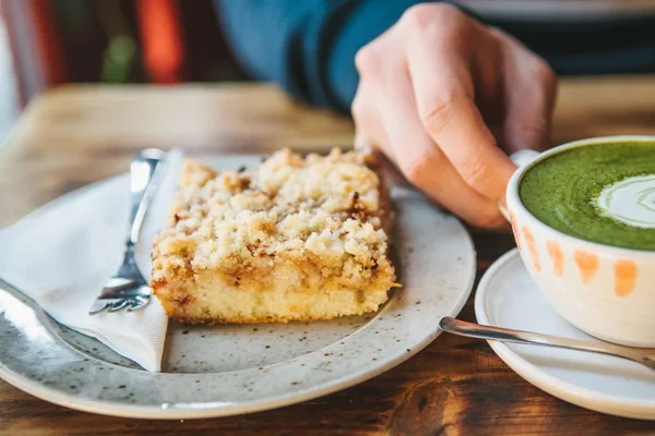 stock image Close-up mans hand holding mug of green tea with beautiful pattern in the form of white foam next to dessert