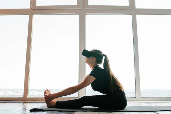 Une jeune belle fille en lunettes de réalité virtuelle fait du yoga et de l'aérobic à distance. Concept technologique futur. Technologie d'imagerie moderne. Cours de sport individuel à distance . — Photo