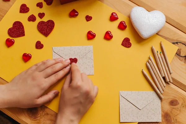 The girl wrote a letter with congratulations and pastes the envelope with a symbol in the form of a heart. Nearby lie various objects symbolizing the event.