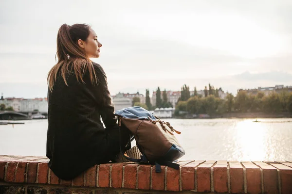 Uma menina turística com uma mochila está sentada ao lado do rio no fundo das casas em Praga, na República Checa, na Europa. Viagens, lazer, turismo . — Fotografia de Stock