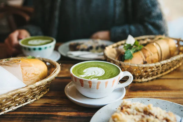 Девушка сидит в кафе и держит кружку с зеленым чаем с молоком Маття рядом с кусочком сладкого пирога и двумя сэндвичами с овощами. Завтрак в кафе . — стоковое фото