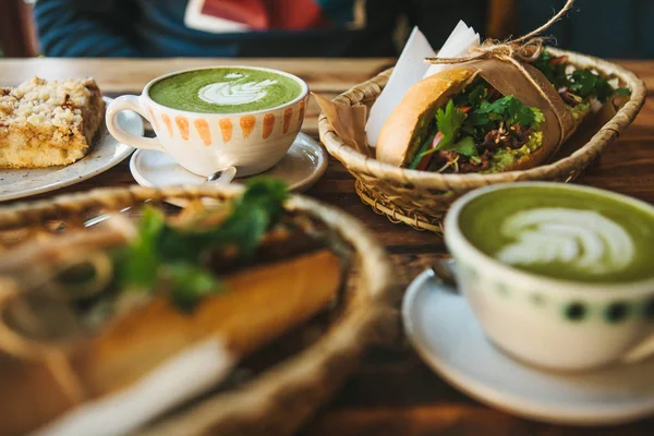 Много разных блюд на столе. Здоровый завтрак в ресторане: чашки зеленого чая с молоком Matcha, десерт и сэндвичи с овощами и травами на деревянном столе . — стоковое фото