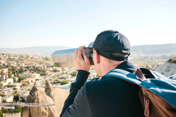 A traveler with a backpack on at viewpoint looks through binoculars at a beautiful view of the town of Goreme in Cappadocia in Turkey. Journey. Hiking.