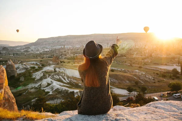 Een toeristische meisje in een hoed zit op een berg en kijkt naar de zonsopgang en ballonnen in Cappadocië. Toerisme, attracties, Turkije. — Stockfoto