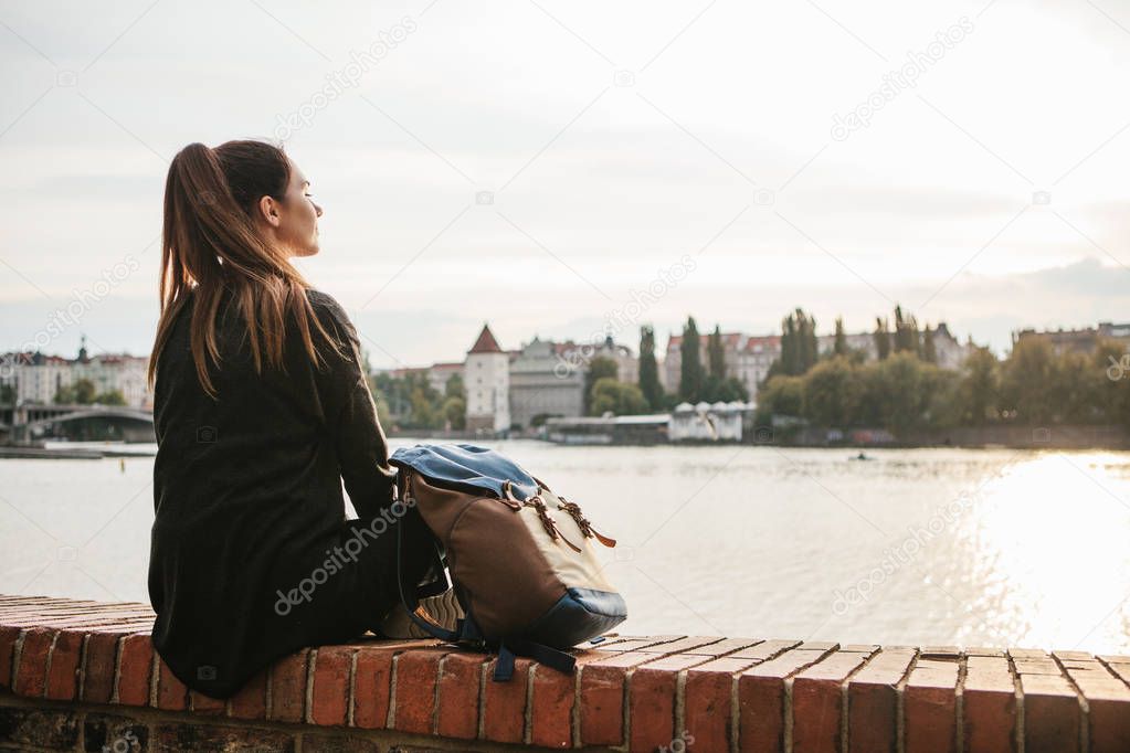 A tourist girl with a backpack is sitting next to the river on the background of houses in Prague in the Czech Republic in Europe. Travel, leisure, tourism.