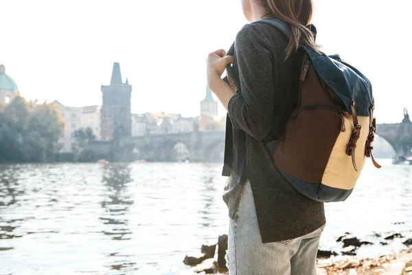 Una hermosa joven turista con una mochila se encuentra junto al río Moldava en Praga y admira una de las atracciones más populares llamado el Puente de Carlos. Viajar por la República Checa . — Foto de Stock