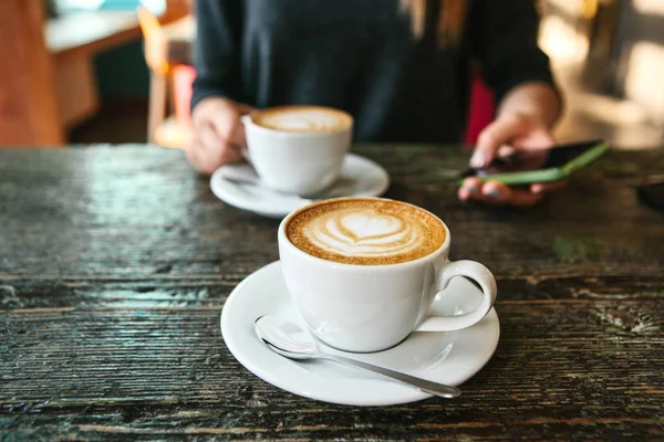 Two cups of coffee on a wooden table, a young girl holding a phone in her hand and going to call. Waiting for a meeting. Another person is late for the meeting.