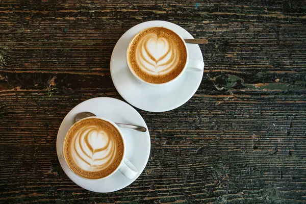 Vista superior. Dos tazas de delicioso café cappuccino fragante con espuma y una amasadora sobre una superficie de madera . — Foto de Stock
