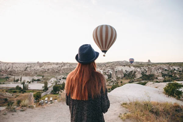 A tourist girl in a hat admires hot air balloons flying in the sky over Cappadocia in Turkey. Impressive sight. — Stock Photo, Image