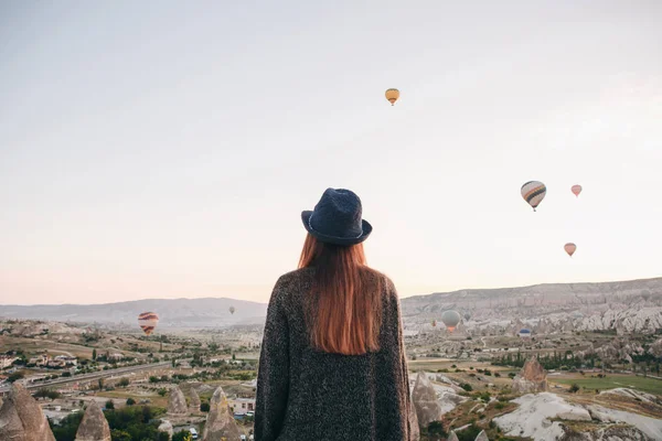 A tourist girl in a hat admires hot air balloons flying in the sky over Cappadocia in Turkey. Impressive sight. — Stock Photo, Image