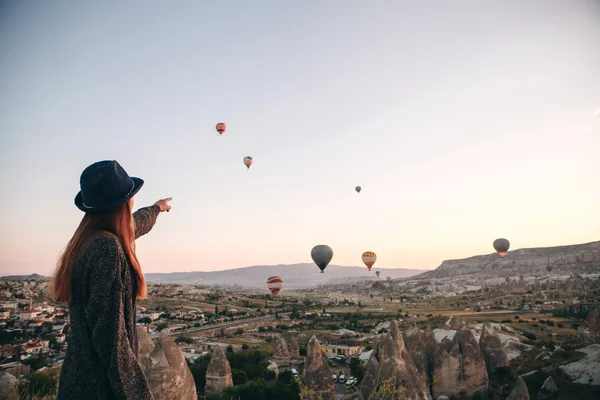 O fată turistică într-o pălărie admiră baloanele cu aer cald care zboară pe cer deasupra Cappadociei din Turcia. Vedere impresionantă . — Fotografie, imagine de stoc