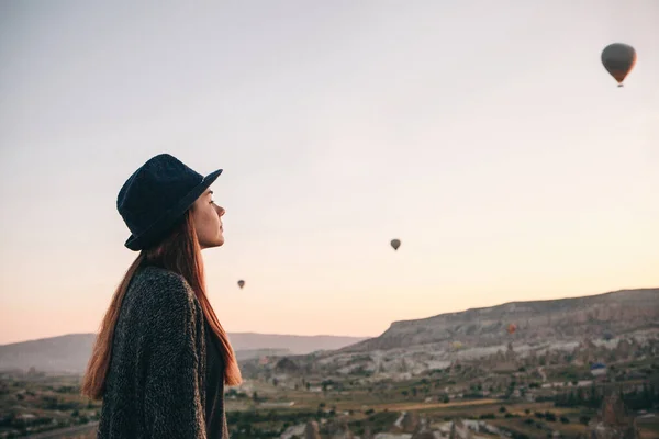 A tourist girl in a hat admires hot air balloons flying in the sky over Cappadocia in Turkey. Impressive sight. — Stock Photo, Image