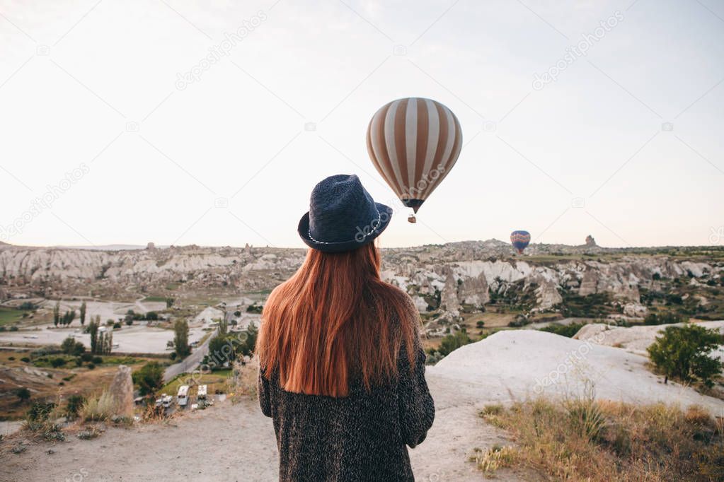 A tourist girl in a hat admires hot air balloons flying in the sky over Cappadocia in Turkey. Impressive sight.