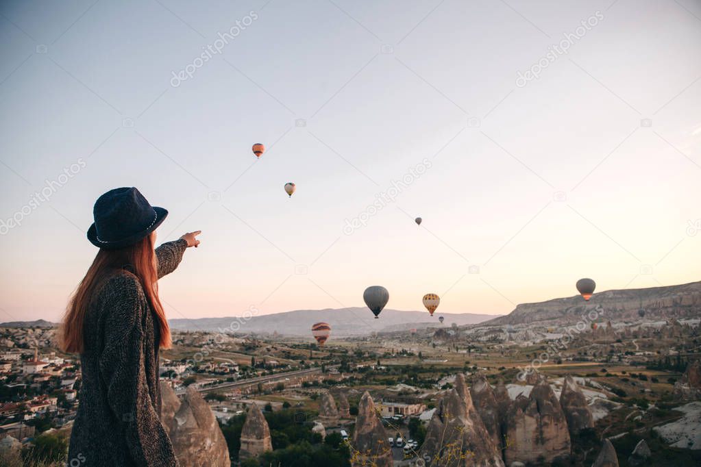 A tourist girl in a hat admires hot air balloons flying in the sky over Cappadocia in Turkey. Impressive sight.