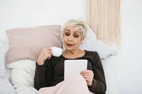 Una anciana acostada en la cama bebiendo café de la mañana usa una tableta para ver noticias o charlar con amigos en una red social. Generación de personas mayores y nuevas tecnologías . — Foto de Stock