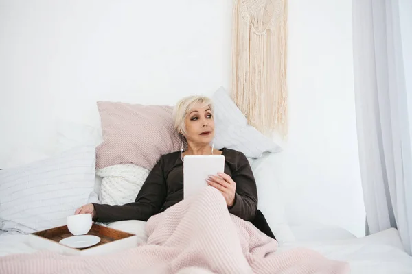 Una anciana acostada en la cama bebiendo café de la mañana usa una tableta para ver noticias o charlar con amigos en una red social. Generación de personas mayores y nuevas tecnologías . — Foto de Stock