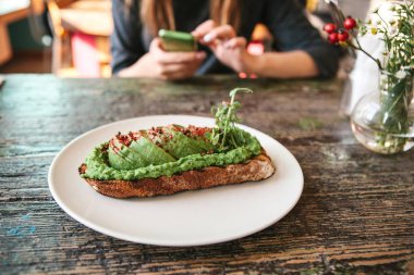 Ahşap bir masa üzerinde avacado ile sandviç. Kız arka planda başka bir kişiye bir ileti yazın veya aramak için gidiyor.