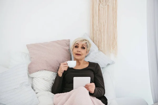 Una anciana acostada en la cama bebiendo café de la mañana usa una tableta para ver noticias o charlar con amigos en una red social. Generación de personas mayores y nuevas tecnologías . — Foto de Stock