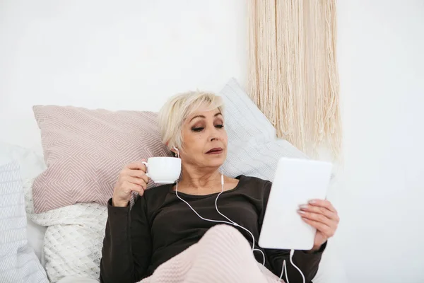 Una mujer positiva bebe un café de la mañana acostada en la cama y usa una tableta para ver videos, escuchar música y charlar con amigos en las redes sociales . — Foto de Stock