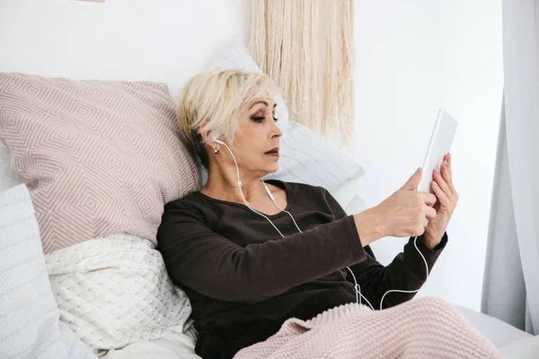 Una mujer mayor positiva utiliza una tableta para ver videos, escuchar música y chatear con amigos en las redes sociales . — Foto de Stock