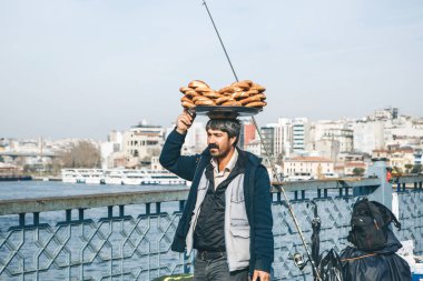 Türkiye, İstanbul, 29 Aralık 2019 - Satıcı Galata köprüsü boyunca yürüyor ve kafasında çok sayıda geleneksel Türk simidi taşıyor. Sokak yemekleri.