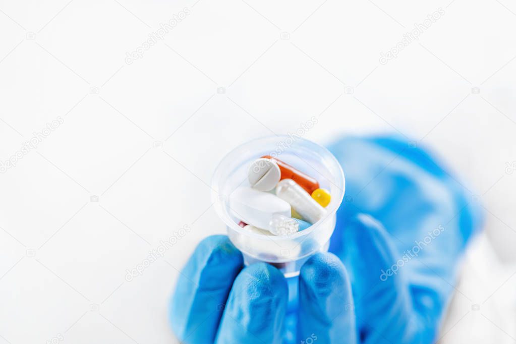 Female doctor`s hand In blue glove holding graduated medicine cups full of pills. Copy space and white background.