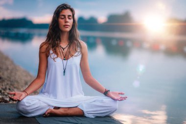 Güzel genç bir kadın göl kenarında yoga egzersizi yapıyor. Lotus pozisyonunda oturuyor.