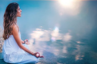 Güzel genç bir kadın göl kenarında yoga egzersizi yapıyor. Lotus pozisyonunda oturuyor.