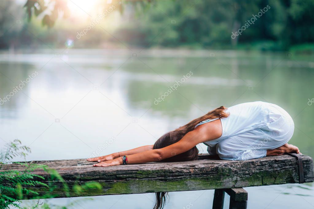 Woman meditates in Balasana, child's pose by the lake