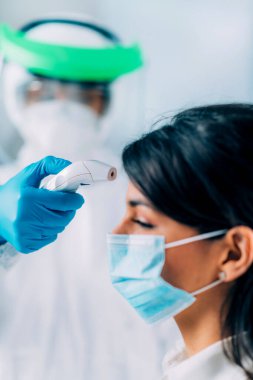 Coronavirus taraması. Koruyucu kıyafetli, vücut ısısını ölçen, temassız vücut termometresi olan, koruyucu maske takan genç bir kadın.