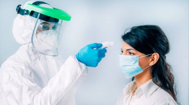 Coronavirus taraması. Koruyucu kıyafetli, vücut ısısını ölçen, temassız vücut termometresi olan, koruyucu maske takan genç bir kadın.