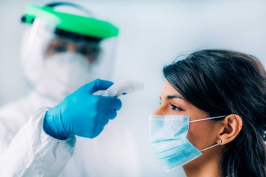 Coronavirus taraması. Koruyucu kıyafetli, vücut ısısını ölçen, temassız vücut termometresi olan, koruyucu maske takan genç bir kadın.