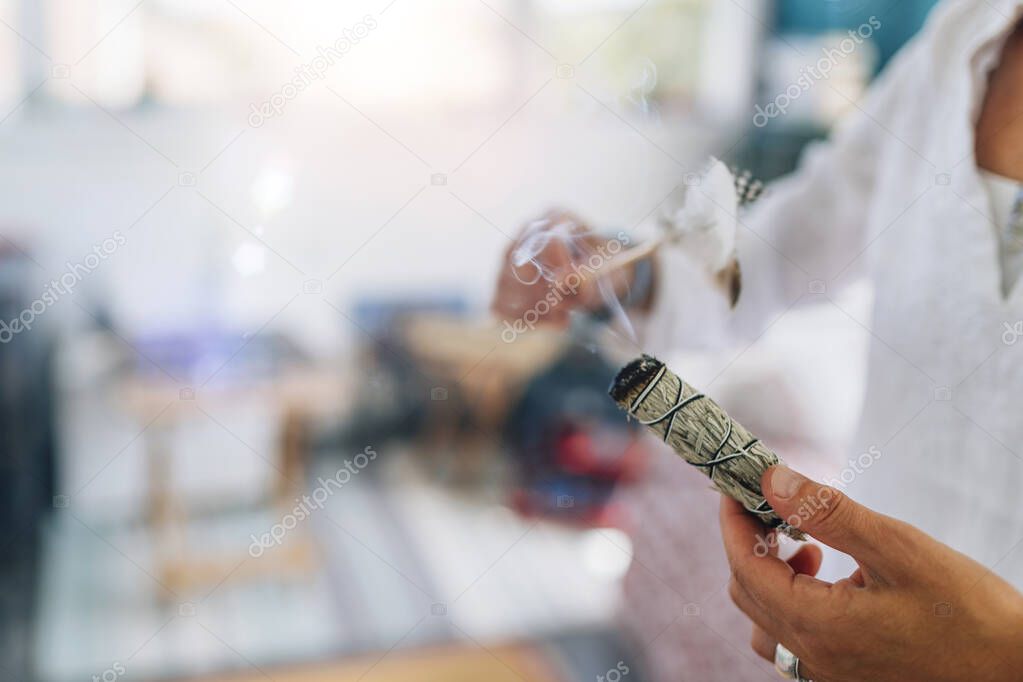 Womans hand holding burning sage smudge stick, spreading the smoke around with white feather, blessing and cleansing home from negative energies 